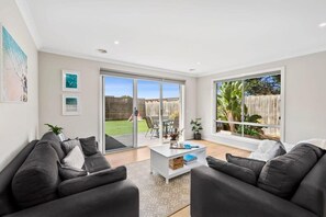 A modern living room with a sofa set, a coffee table, and large windows offering a view of the outdoors. - Seahaze Retreat | Spacious Family Coastal Haven (Torquay)