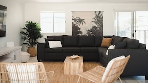 Un salon moderne avec un canapé d’angle noir, une table basse en bois et une grande fresque murale aux motifs tropicaux.