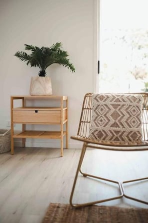 A room with a wooden side table, a potted plant, a wicker chair, and a window. - Seagrass (Culburra Beach)