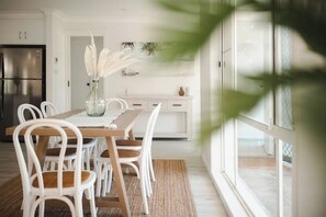 A modern kitchen with white cabinetry, a stainless steel refrigerator, and a dining area with a wooden table and white chairs. - Seagrass (Culburra Beach)