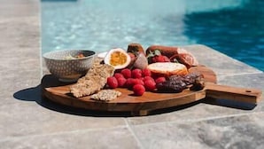 Une scène au bord de la piscine, avec une planche en bois sur laquelle sont disposés des en-cas et un saladier, et en arrière-plan, une maison moderne avec une façade blanche et un toit noir.
