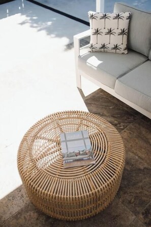 Un pouf rond en osier, sur lequel repose un magazine, est placé sur un sol en béton, à côté d’un canapé blanc orné d’un coussin à motifs de palmiers.