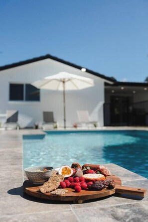 Un espace aménagé au bord de la piscine, avec une planche en bois servant de présentoir pour des collations et un parasol blanc.