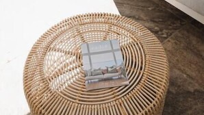 Un pouf rond en osier, surmonté d’une petite boîte, est placé sur un sol carrelé, près d’un canapé blanc agrémenté d’un coussin à motifs de palmiers.