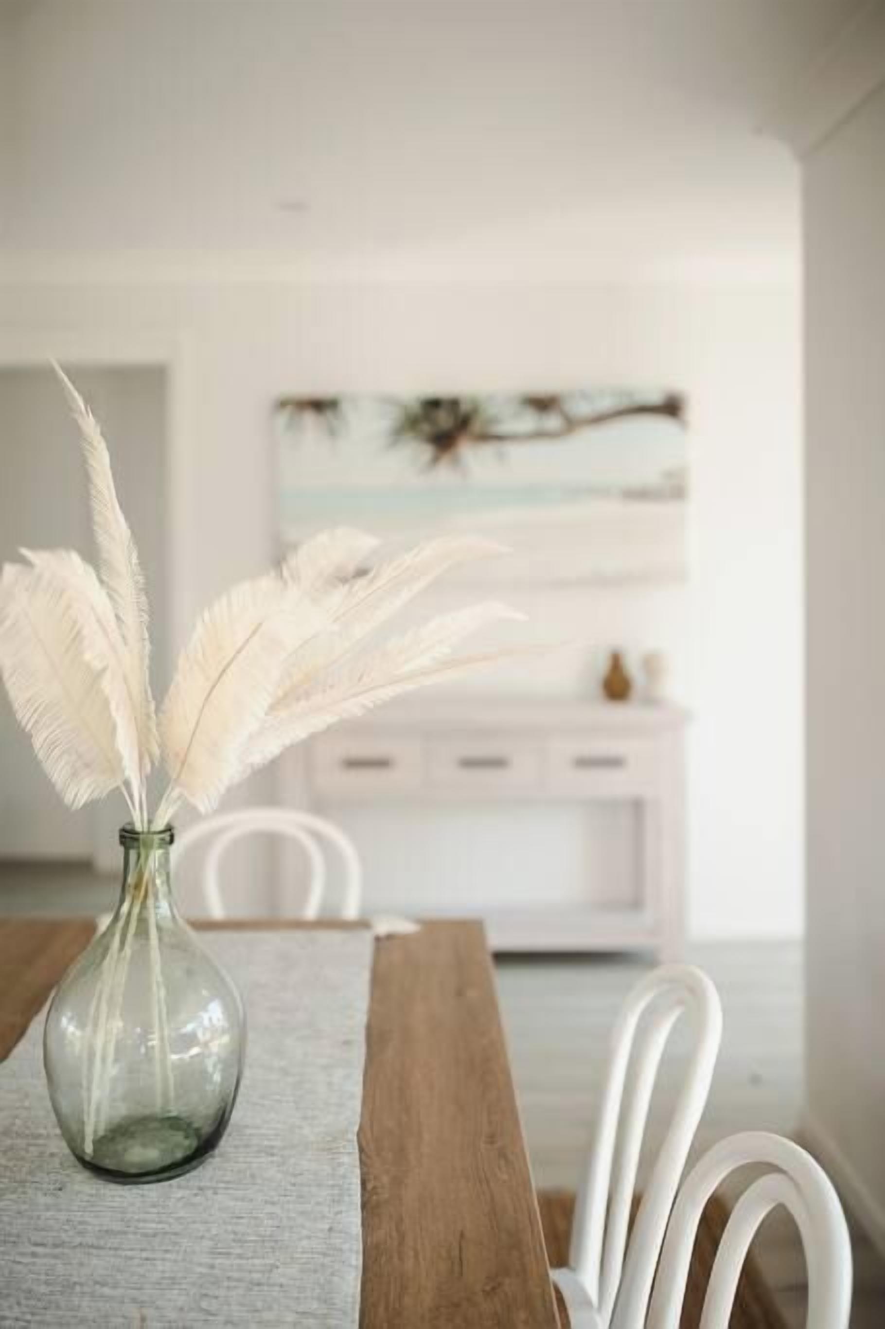 Una mesa de comedor con un florero de plumas blancas, camino de mesa de madera y sillas blancas.