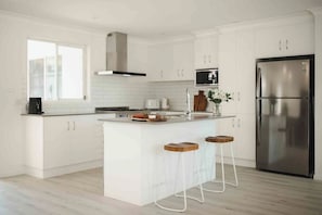 A modern kitchen with white cabinetry, a stainless steel refrigerator, and a central island with stools. - Seagrass (Culburra Beach)