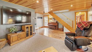 Amplia sala de estar con techo de madera, un televisor de pantalla plana grande, un piano y un sillón reclinable.