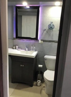 Une petite salle de bain avec un lavabo blanc, des meubles sombres, des toilettes et un miroir doté d’un éclairage violet.