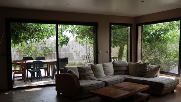 A modern living room with a sectional sofa, a wooden coffee table, and a dining area with chairs and a table. Large windows provide a view of a garden. - A break for two in a detached villa with SPA (Sainte-Anastasie)