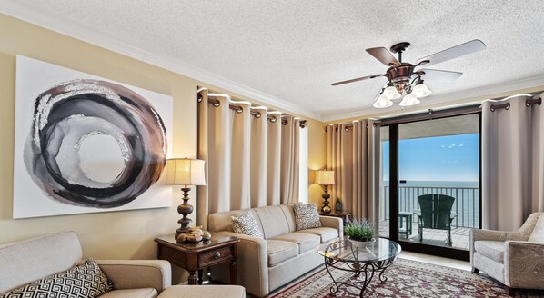 Quarto de hotel com uma grande pintura abstrata, um ventilador de teto, um sofá, uma mesa de centro e vista para o oceano através de uma porta deslizante.