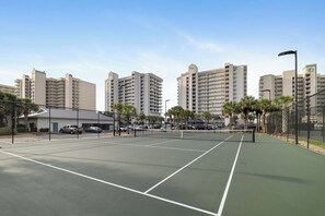 Una cancha de tenis con una red, rodeada por una valla, y edificios altos al fondo.