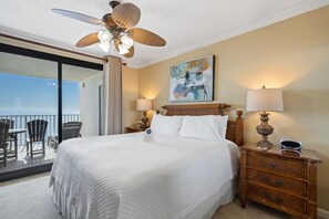 Quarto de hotel com uma cama grande, duas mesas de cabeceira com abajures, um ventilador de teto, um quadro na parede e uma varanda com cadeiras.