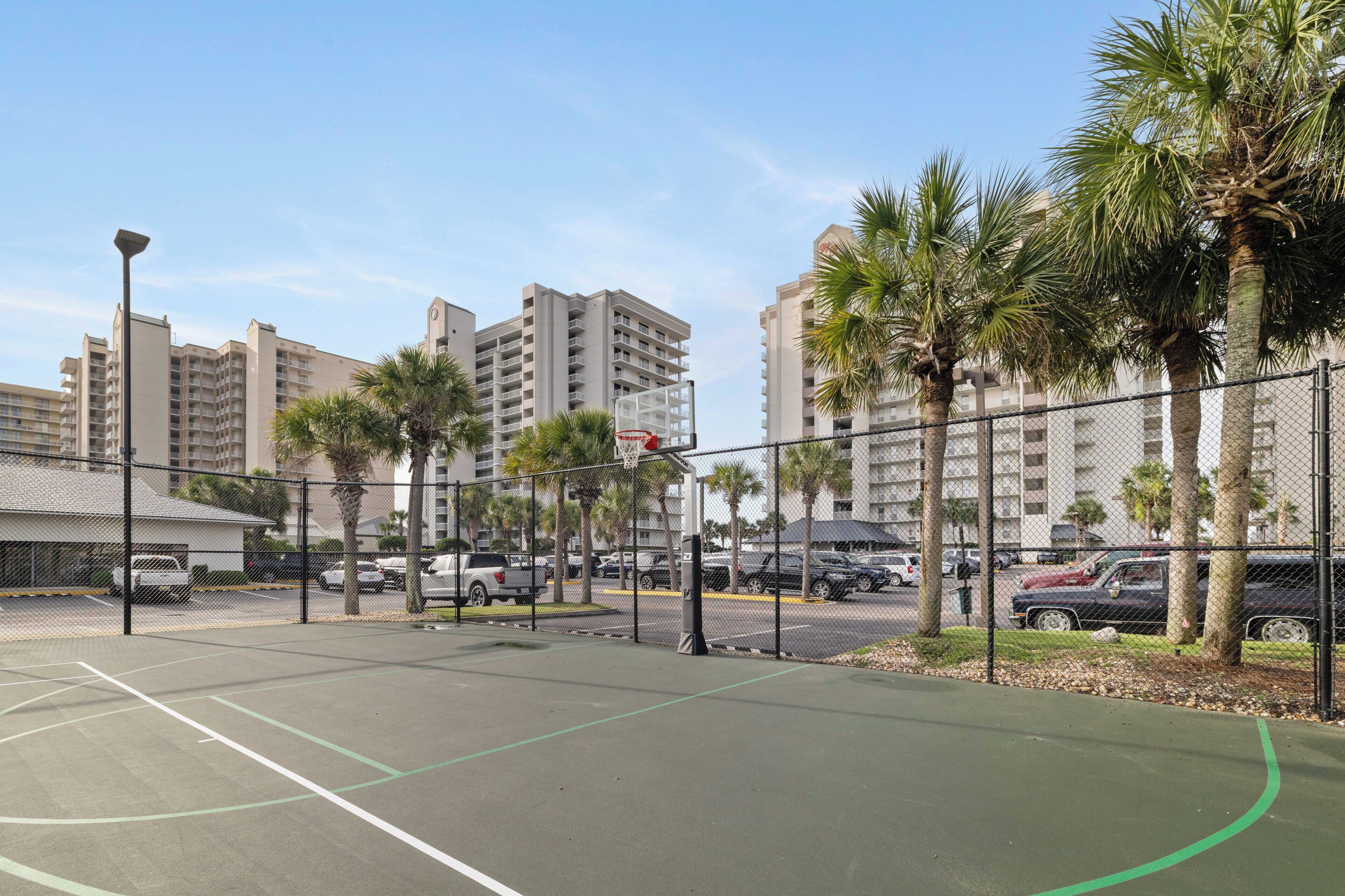 A tennis court surrounded by a fence, with palm trees and a parking lot in the background.