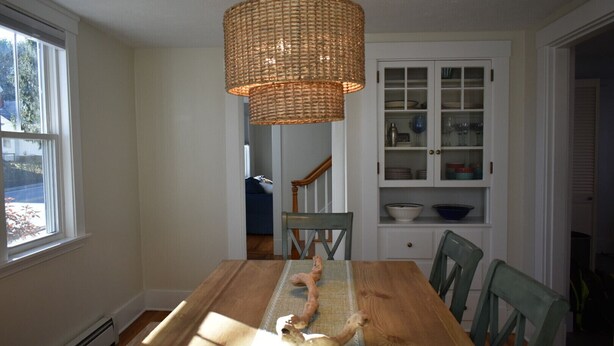 Un coin repas avec une table en bois, un luminaire suspendu en osier et une vue sur une cuisine équipée d’armoires et d’un escalier.