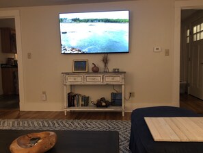 A living room with a television mounted on the wall, a bookshelf with books and decorative items, a wooden bench, and a patterned rug.