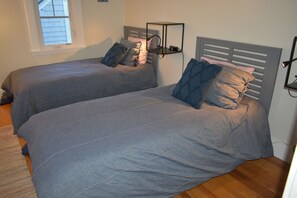A bedroom with two beds, each with a headboard and pillows. There is a window above the beds, and a shelf with a lamp on the wall.