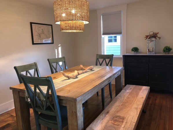 A dining area with a wooden table, four chairs, a bench, a woven pendant light, a framed map, a window with blinds, and a small plant on a cabinet.