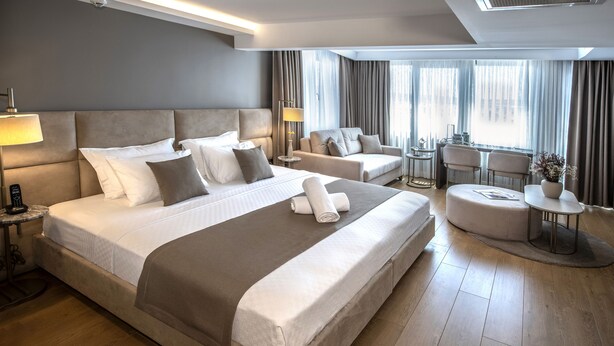 A modern hotel room with a large bed, a sofa, a small table, and a view of the city through large windows. - Valente Suites & Hotel (Istanbul)