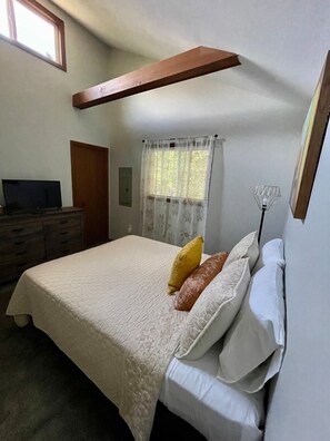 A bedroom with a bed, a dresser, a television, a window with curtains, and a ceiling beam.