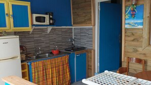 A compact kitchen with a microwave, sink, and a small refrigerator. There is a wooden table and a chair in the foreground. - Studio eliconia<br>Quartier anse bellune <br>Rue la bellune Trinité 97220 (trinite)