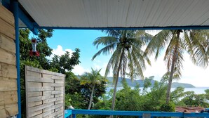 A covered outdoor seating area with a table and chairs, overlooking a scenic view of palm trees and a body of water. - Studio eliconia<br>Quartier anse bellune <br>Rue la bellune Trinité 97220 (trinite)