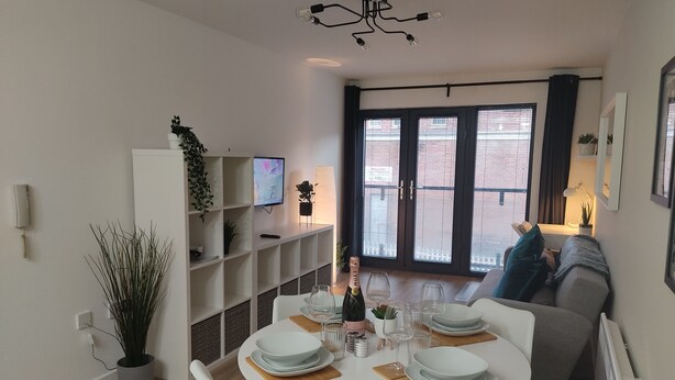 A modern dining area with a round table set for four, a TV mounted on the wall, and a balcony with a view of buildings. - Fantastic Centrally Located 1 bed Apartment (Liverpool)