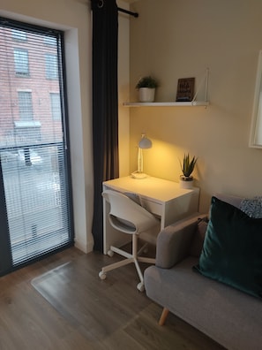 Ein modernes Zimmer mit weißem Schreibtisch, grauem Bürostuhl, grauem Sofa mit grünem Kissen, einem Topf mit Pflanze und einem Regal mit einem gerahmten Bild.