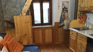 A cozy room with a wooden floor, a blue sofa with orange cushions, a kitchen area with wooden cabinets and a sink, and a window with a view of greenery. - Monterosa winter vacation home (Gressonay Saint Jean)