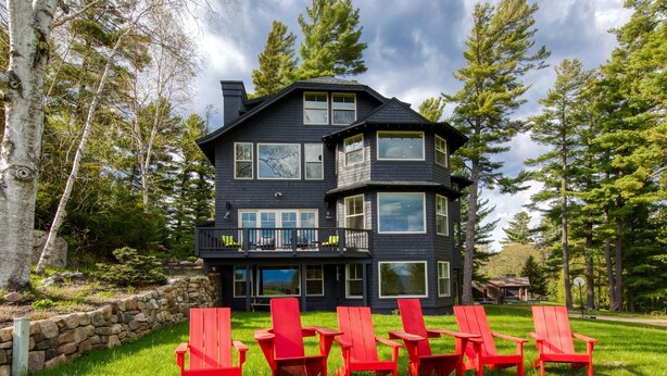 A two-story house with a balcony, surrounded by a stone wall and red chairs in the yard.