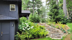 A house with a dark exterior, a stone staircase leading to a wooden deck, surrounded by lush greenery and tall trees.