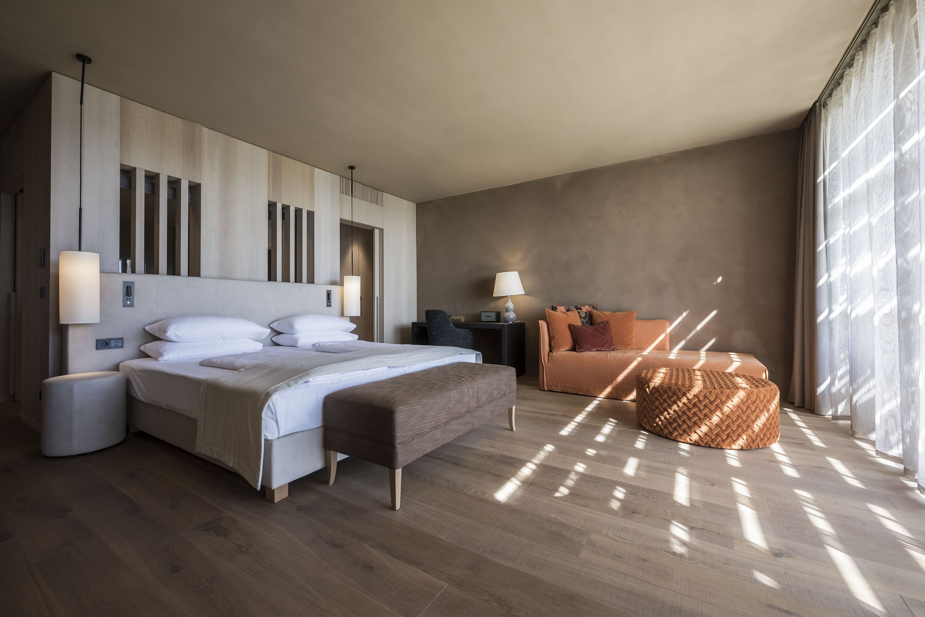 A modern hotel room with a large bed, a sofa, a round ottoman, and a nightstand.