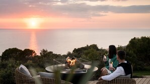 A couple sitting by a fire pit overlooking a sunset over the ocean, with lush greenery in the background.