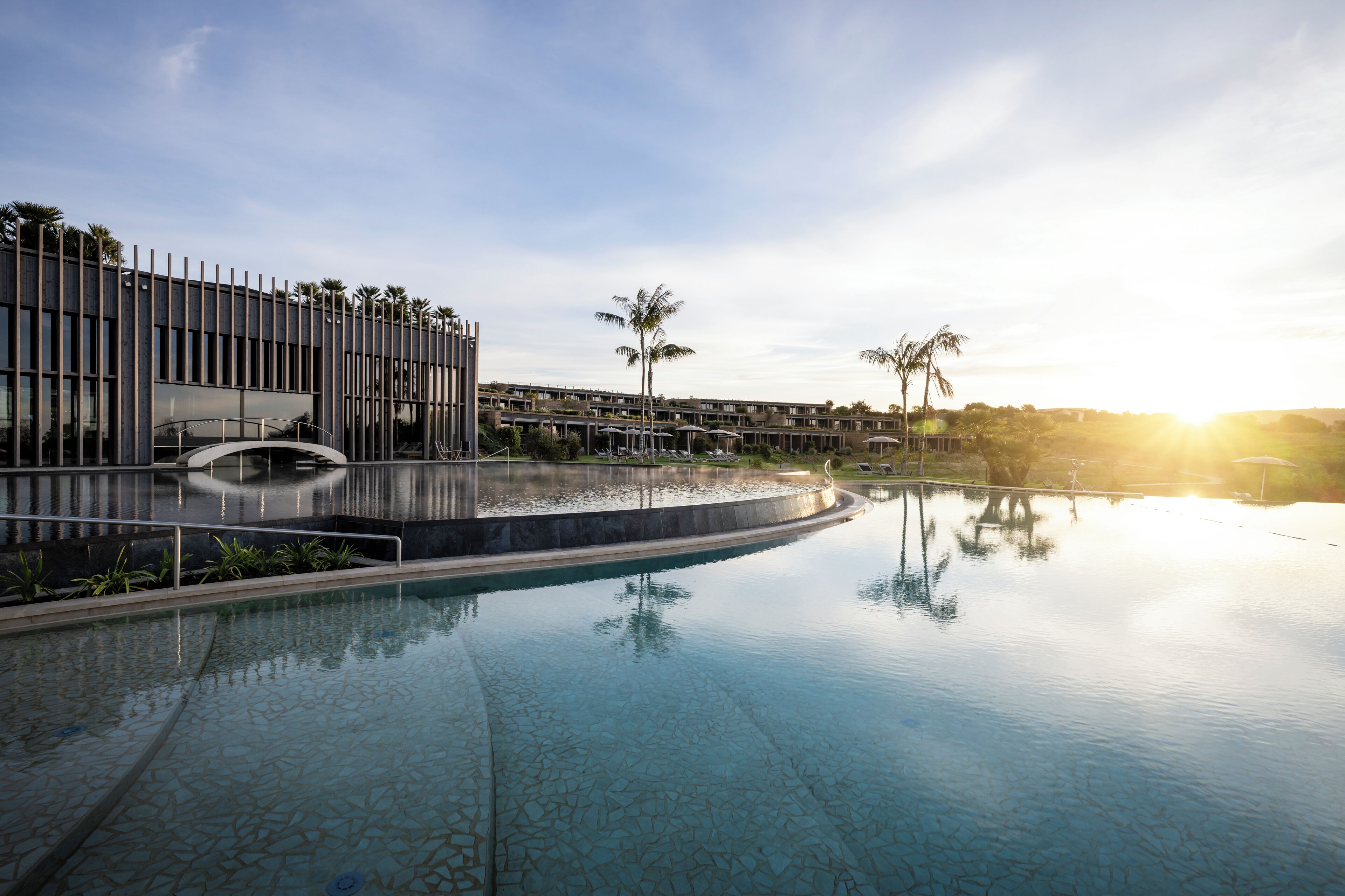 A modern hotel with a swimming pool, palm trees, and a clear sky.