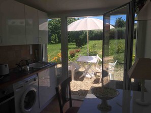 A compact kitchen with a washing machine, a dining area with a table and chairs, and a view of a garden with an umbrella.