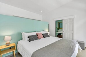 A modern bedroom with a large bed, a bedside table with a lamp, and a door leading to a bathroom. - Home with the best pool 5 min drive to town (tamarindo)