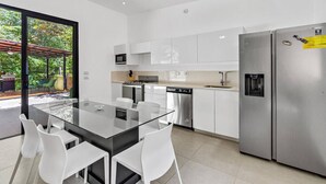 A modern kitchen with a glass dining table, white chairs, stainless steel appliances, and a view of a garden through large windows. - Home with the best pool 5 min drive to town (tamarindo)