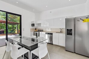 A modern kitchen with a glass dining table, white chairs, stainless steel appliances, and a view of a garden through large windows. - Home with the best pool 5 min drive to town (tamarindo)