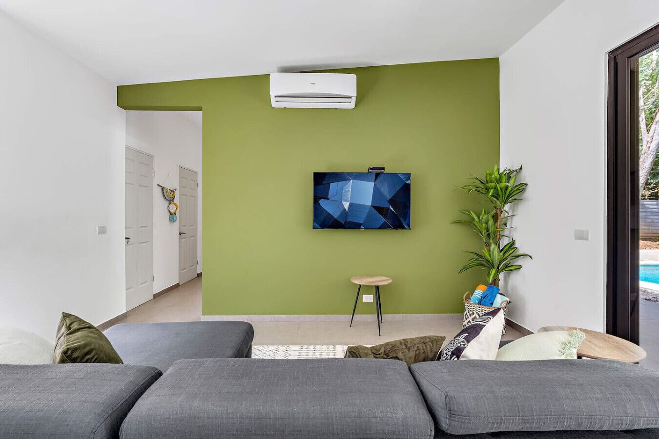 Un salon moderne avec un mur d’accent vert, un téléviseur fixé au mur, une plante en pot et un canapé gris.
