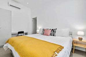 A hotel room with a bed, a desk, a chair, a bedside table with a lamp, and an air conditioning unit on the wall. - Home with the best pool 5 min drive to town (tamarindo)
