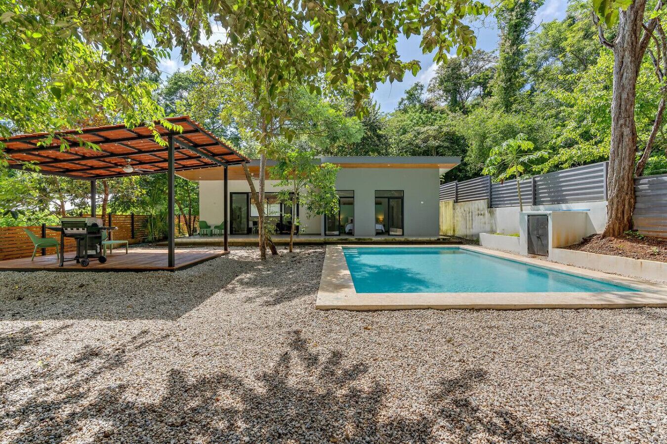 A modern outdoor pool area with a covered patio, a barbecue setup, and a house in the background.