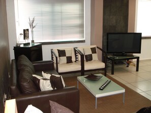 Ein Wohnzimmer mit einer Couch, einem Sessel und einem Couchtisch. Es gibt einen Fernseher und einen kleinen Tisch mit Dekoration.