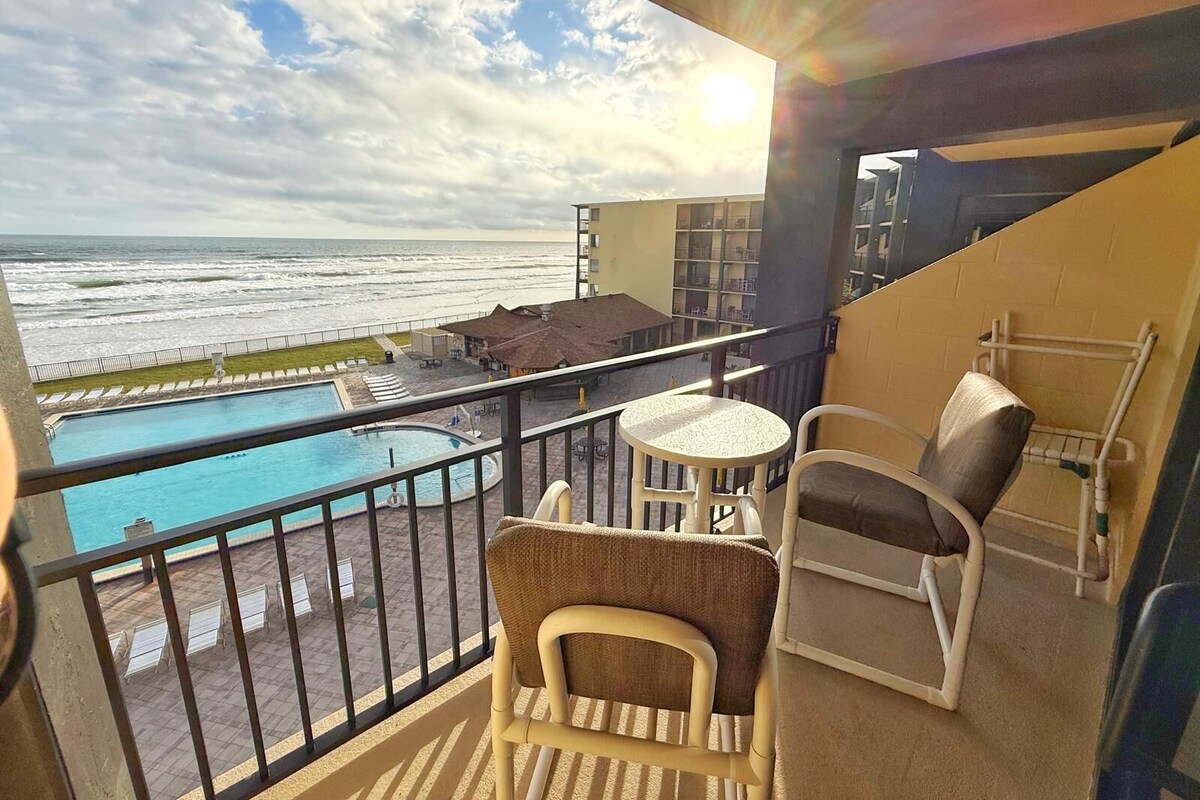 A balcony with a view of the ocean, a pool, and outdoor seating.