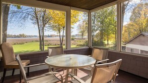 Un patio techado con una mesa redonda y sillas, ventanas grandes que ofrecen vista a árboles y una masa de agua.
