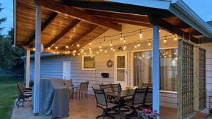 Un patio exterior con zona de estar cubierta, luces colgantes y una parrilla.