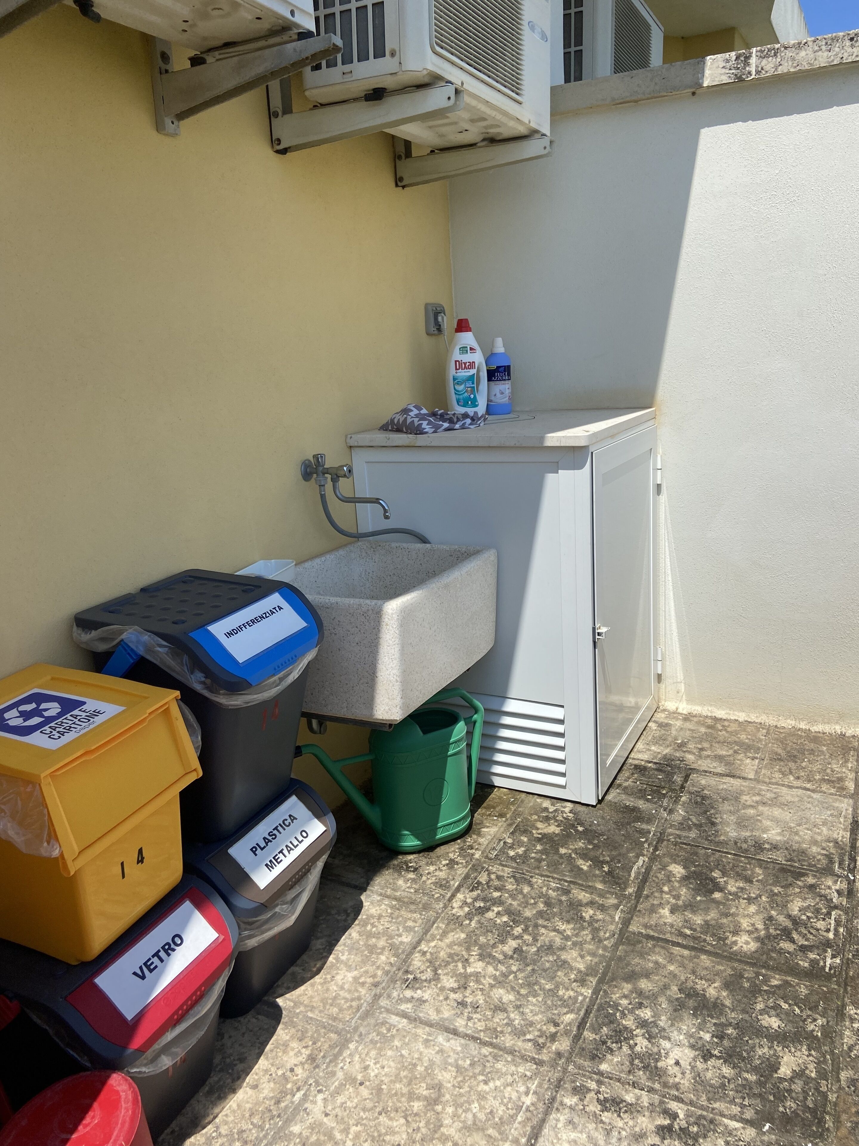 Una kitchenette pequeña con un lavamanos, un refrigerador y un dispensador de agua. Hay botes de basura etiquetados para diferentes tipos de residuos y un regador verde.
