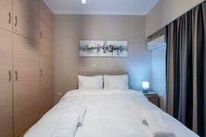 Ein ordentlich bezogenes Bett mit weißen Bettwäsche und Handtüchern, ein großer Kleiderschrank, ein Nachttisch mit Lampe und eine wandmontierte Klimaanlage.