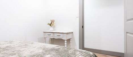 Ein Schlafzimmer mit einem Bett, einer kleinen weißen Kommode mit Schubladen, einer Vase mit Blumen und Holzboden.