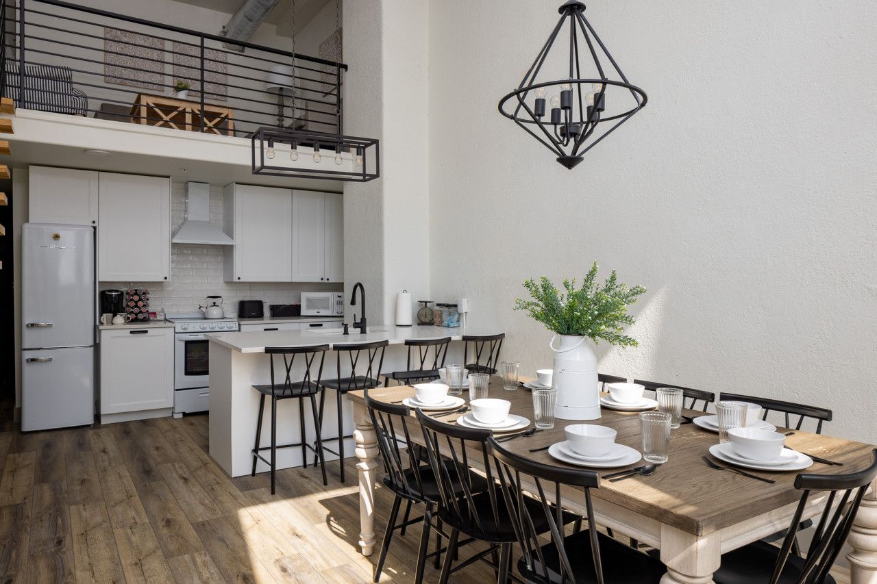A modern kitchen with white cabinets, a dining area with a wooden table and black chairs, and a hanging light fixture.