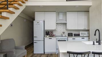 A modern kitchen with white cabinets, a refrigerator, stove, microwave, and a dining area with a white island and bar stools.