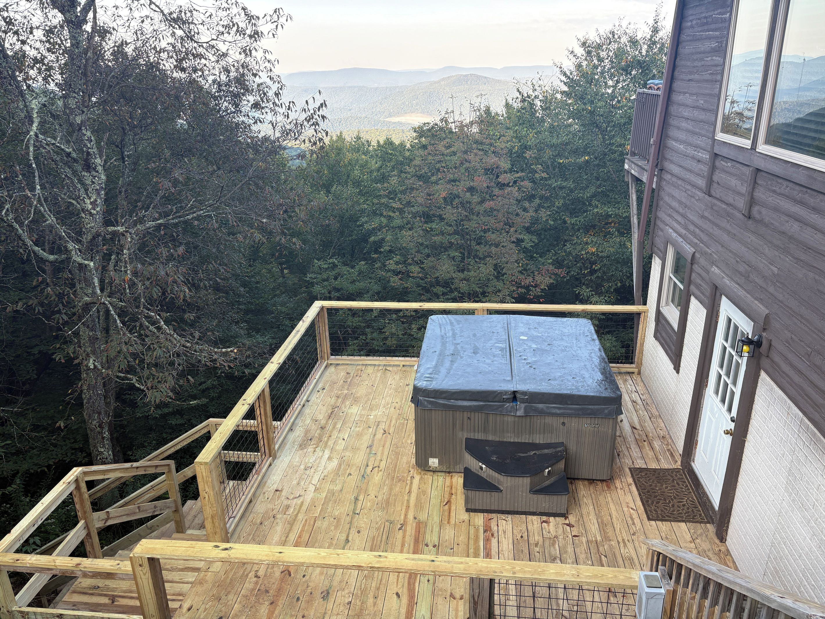 Una terraza de madera con un jacuzzi, rodeada de árboles y vistas a colinas distantes.